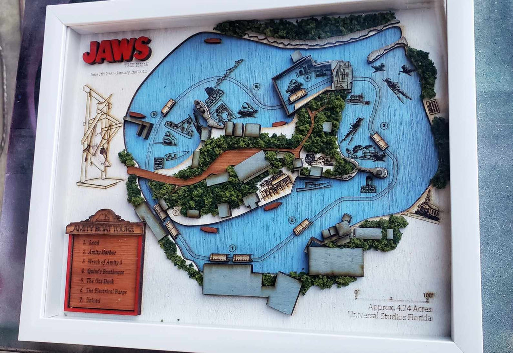 May include: A wooden model of the Jaws ride at Universal Studios Florida. The model is framed in white and features a blue lake with a wooden boat and a shark fin. The model also includes a sign that reads 'JAWS' and 'Approx. 4.74 Acres Universal Studios Florida'. The model also includes a sign that reads 'BOAT TOURS' with a list of tour stops: '1. Lent', '2. Amity Harbor', '3. Wreck of Amity', '4. Squid's Breakfast', '5. Orca'
